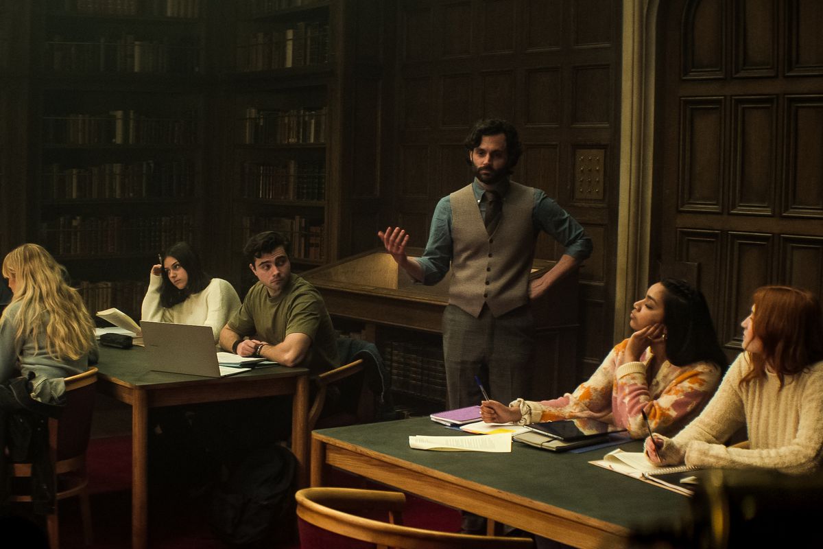 Penn Badgley draagt ​​een vest en praat met een groep studenten in een bibliotheek in You seizoen 4.