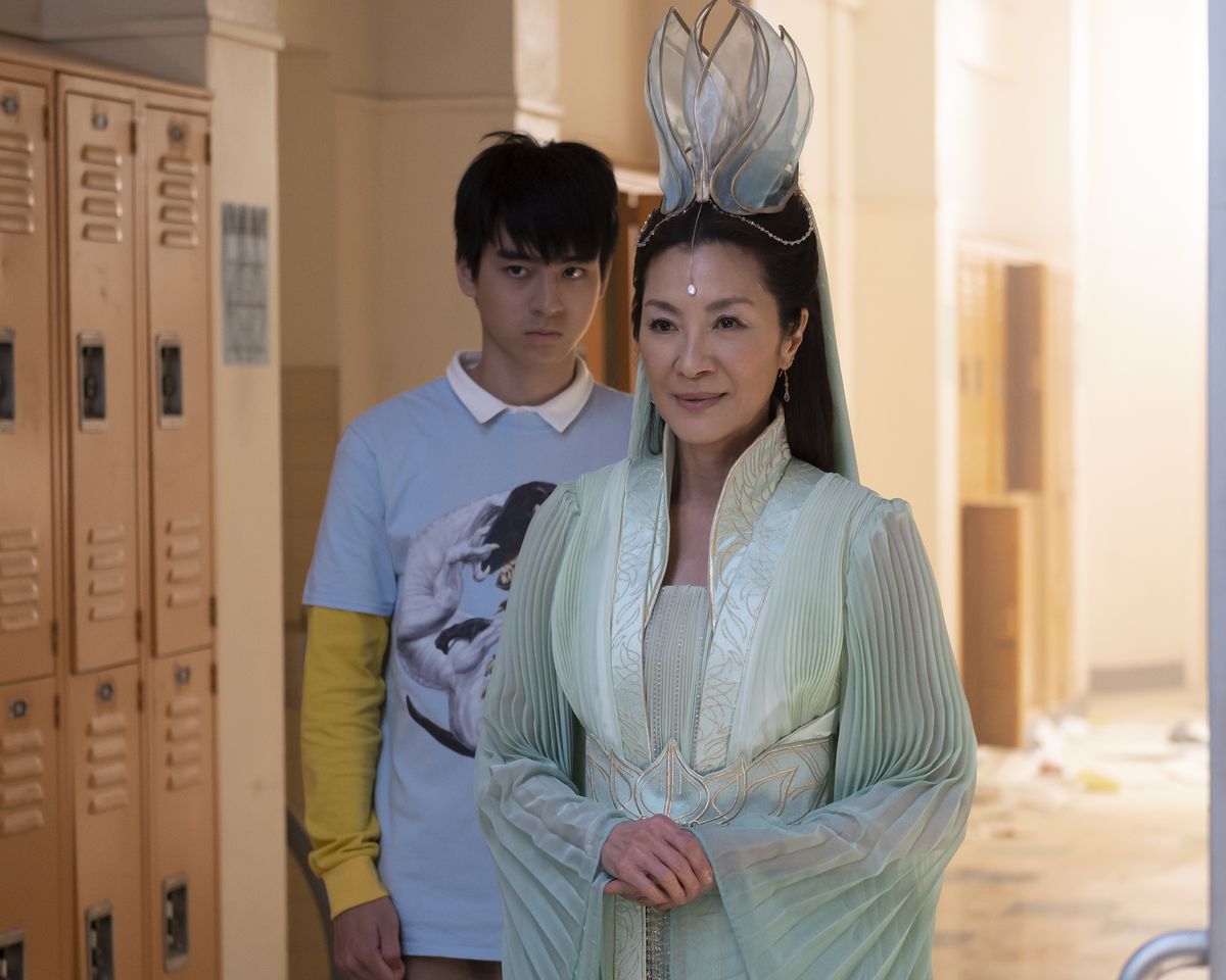 Guanyin (Michelle Yeoh), trong bộ váy kỳ dị, mỉm cười đứng trong hành lang trường học trần tục với những ngăn tủ đựng đồ. Wei-Chen (Jim Liu) đứng sau cô ấy cau mày trong American Born Chinese.