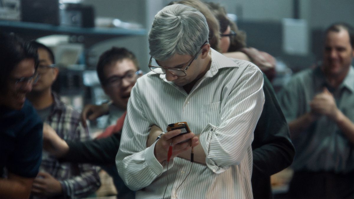Jay Baruchel as a man with graying hair and glasses (Mike Lazaridis) holding a prototype BlackBerry device in BlackBerry.