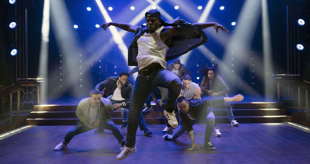Sihirli Mike'ın Son Dansı'ndan bir sahnede, kot pantolon ve ters bir beyzbol şapkası giymiş Siyahi bir erkek dansçı, diğer erkek dansçılar mavi ışıklı bir sahnede onun arkasında çömelirken, parlak beyaz spot ışıklarıyla çaprazlama duruyor.