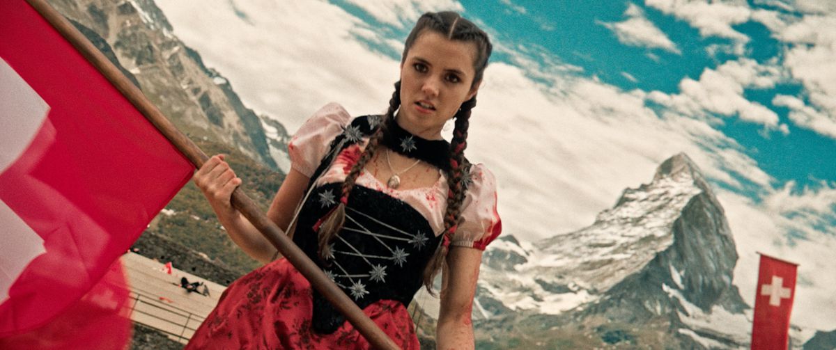 Alice Lucy como Heidi, vestida con un vestido salpicado de sangre, sosteniendo una bandera roja con los Alpes suizos de fondo en Mad Heidi.