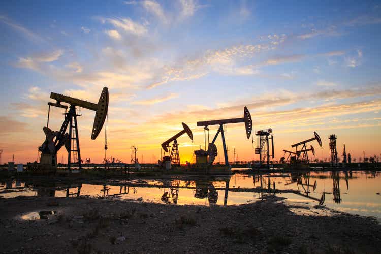 Sitio del campo petrolífero, por la noche, las bombas de petróleo están funcionando, la bomba de petróleo y la hermosa puesta de sol reflejada en el agua