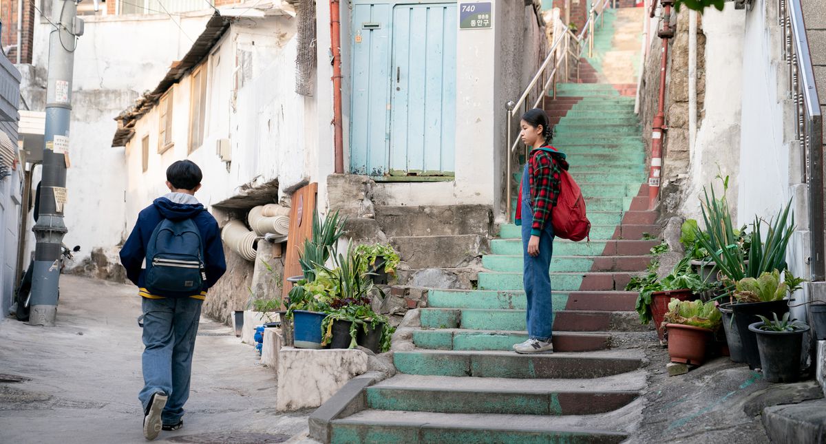 Two children walking home from school. The girl waits on the staircase, watching the boy walk away.