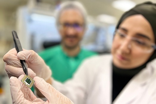 Photo of Esma Ugur, a woman wearing a head covering, lab coat, safety glasses and gloves, holding a chip the size of a small coin. This chip contains the solar cell.