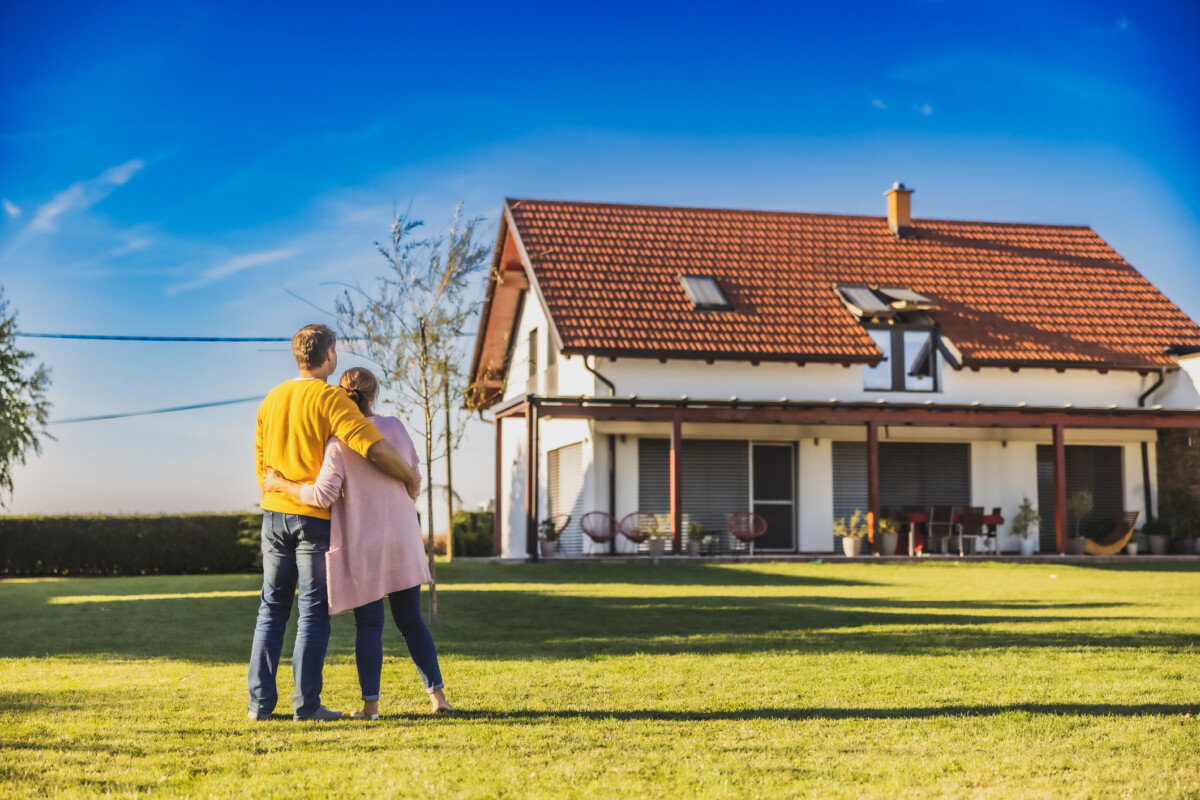 Man en vrouw knuffelen elkaar terwijl ze op het grasveld staan ​​in de achtertuin van hun nieuw gekochte huis, achteraanzicht, modern huis op de achtergrond