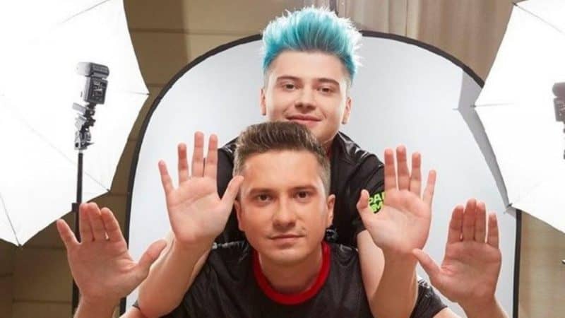 Pro Dota 2 players Roman “RAMZES666” Kushnarev and Alexey “Solo” Berezin pose with their hands up, with one of them above the other. Large white studio lights are in the background.