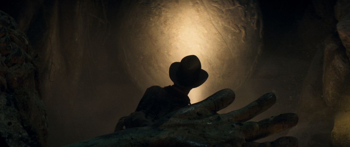Silhouet van Indiana Jones die een tombe verkent, de hand van een lijk is op de voorgrond, ook in de schaduw.