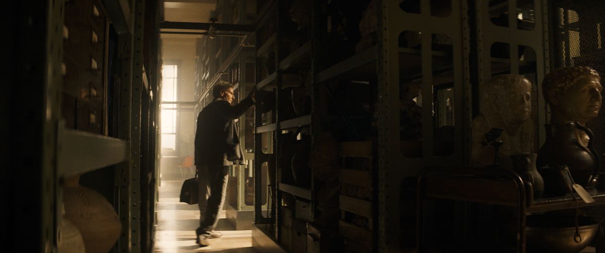 Indiana Jones, gekleed om les te geven, gaat de hoek om tussen grote rekken met boeken en ander materiaal in de archieven van Hunter College.