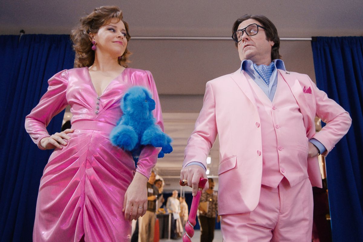 (LR) Elizabeth Banks와 Zach Galifianakis는 The Beanie Bubble에서 파란색 비니 아기와 분홍색 지팡이와 함께 분홍색 정장을 입고 있습니다.