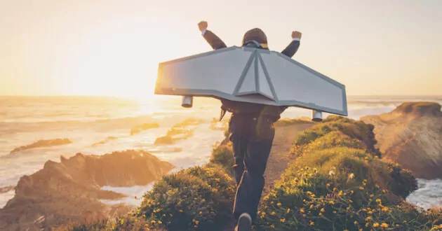 A young boy dressed in business suit and tie wears a homemade jetpack and flying goggles raises his arms in the afternoon sun while running to take off into the air on an outcropping above the surf in Montana de Oro State Park, California. This young entrepreneur is ready to take his new business to new heights.