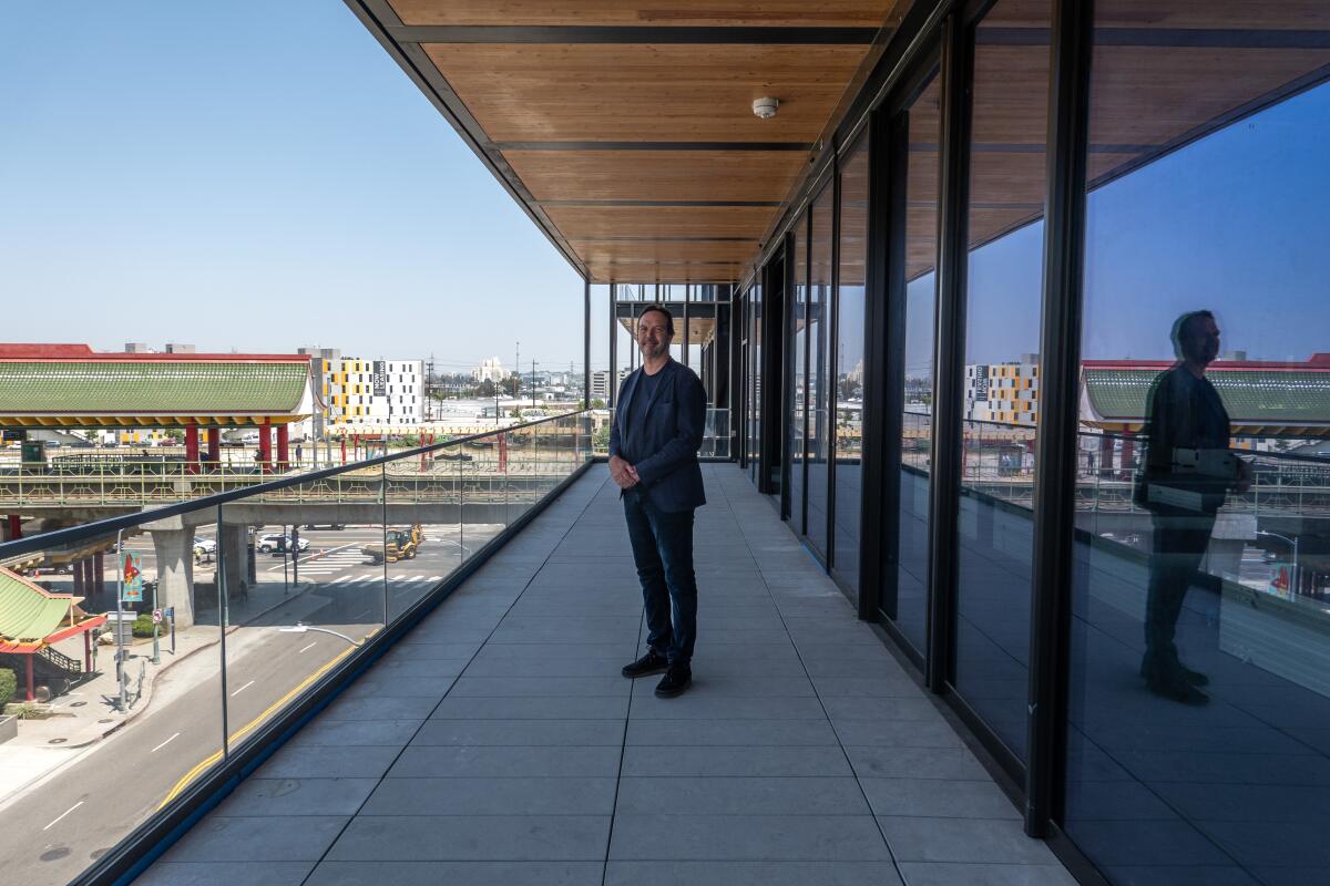 Ein Mann in einem schwarzen Anzug steht auf der Terrasse eines im Bau befindlichen Gebäudes, in der Ferne ist eine erhöhte U-Bahn-Haltestelle zu sehen.