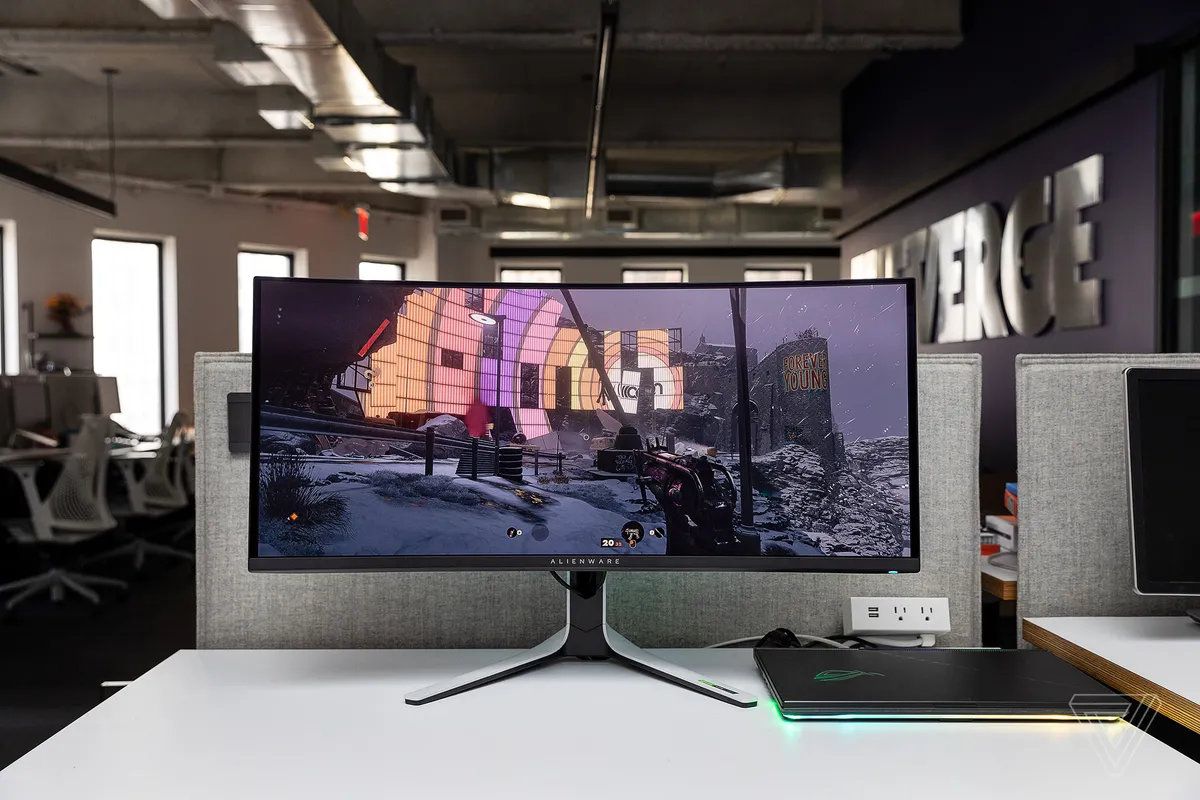 白い机の上に置かれた Alienware AW3423DW 曲面ゲーミング モニターがゲーミング ラップトップに接続されている画像。