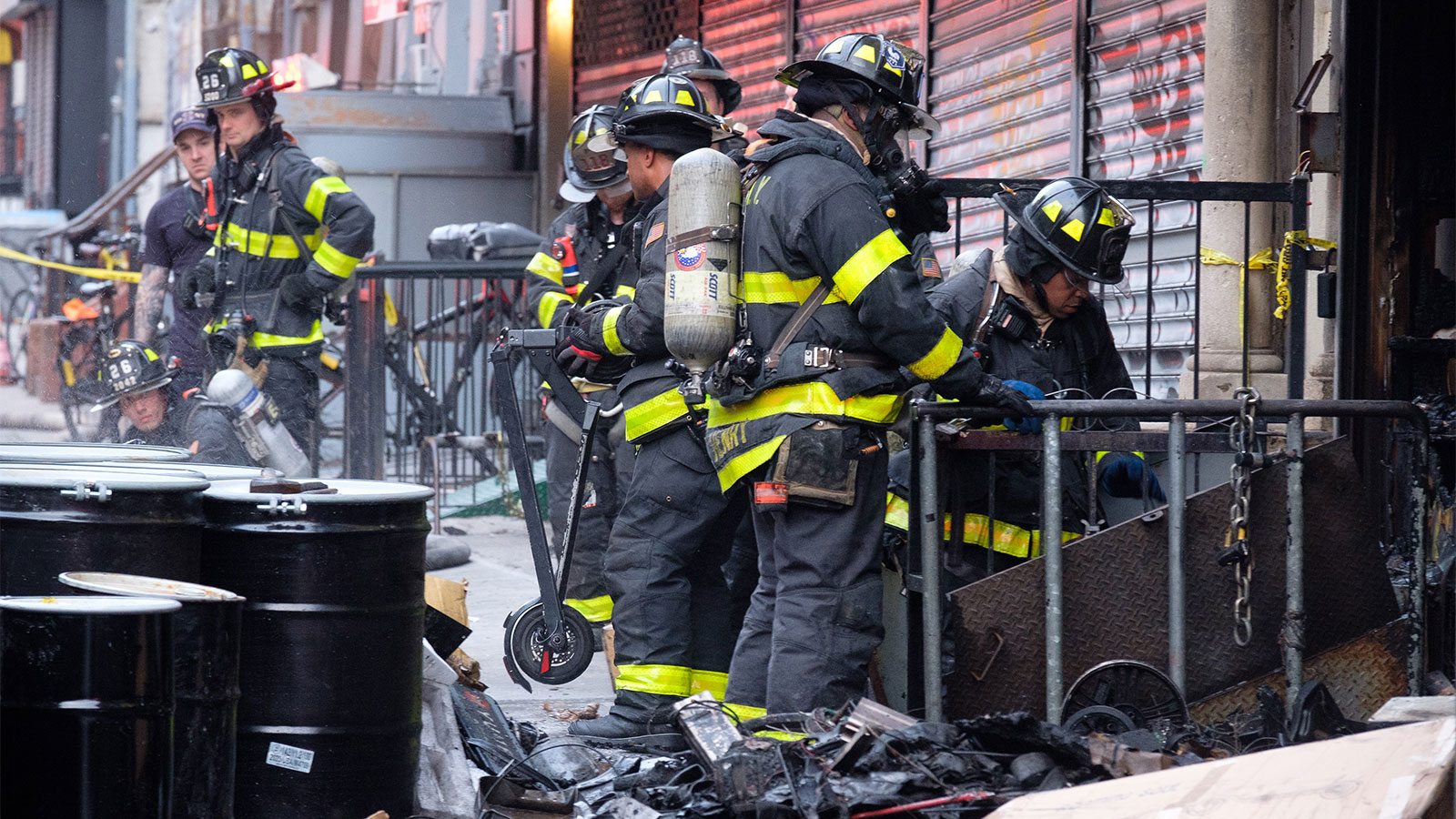 Vários bombeiros usando capacetes e equipamentos pretos à prova de fogo estão em uma calçada entre uma loja fechada, algum lixo e algumas lixeiras cilíndricas