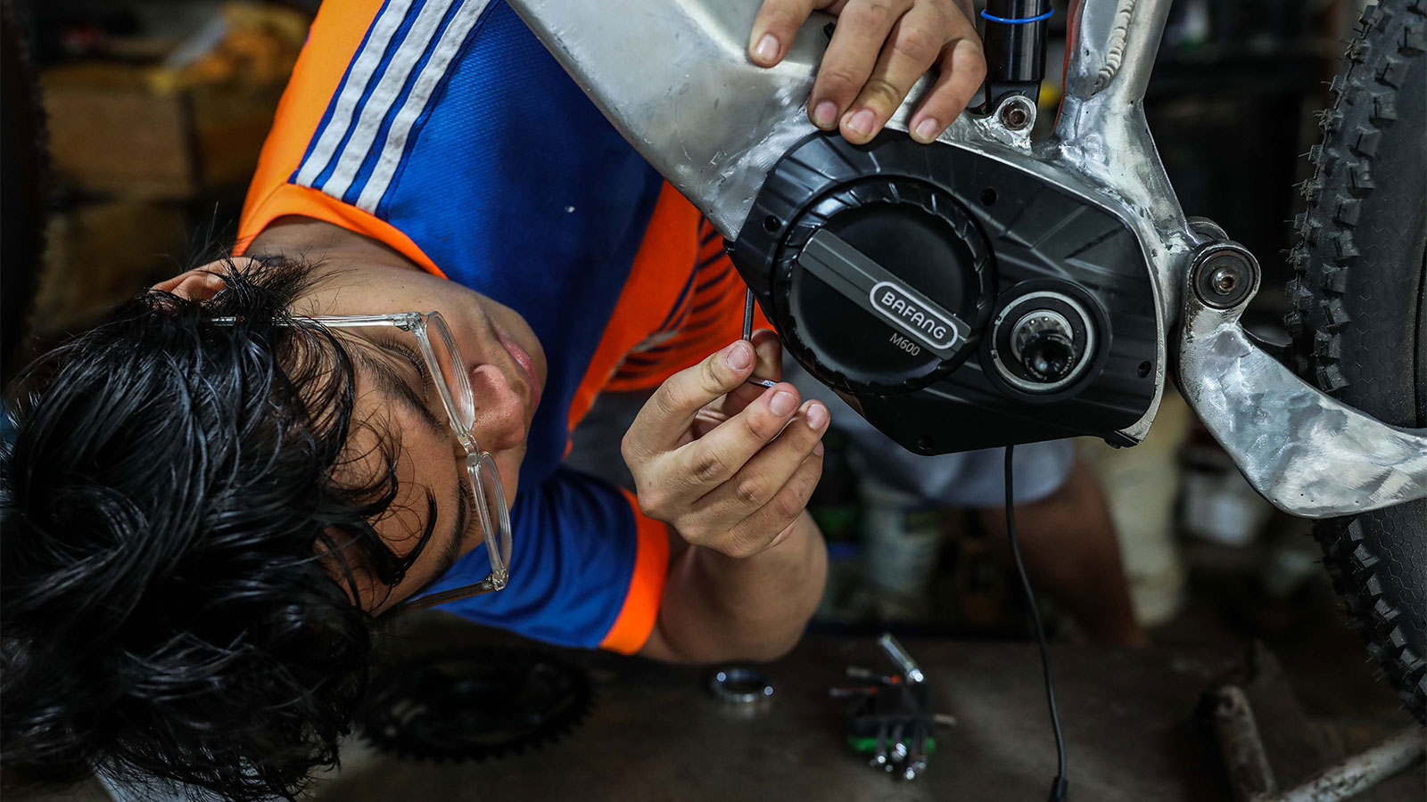 Um homem de óculos se inclina para trabalhar no quadro de uma bicicleta elétrica com uma ferramenta