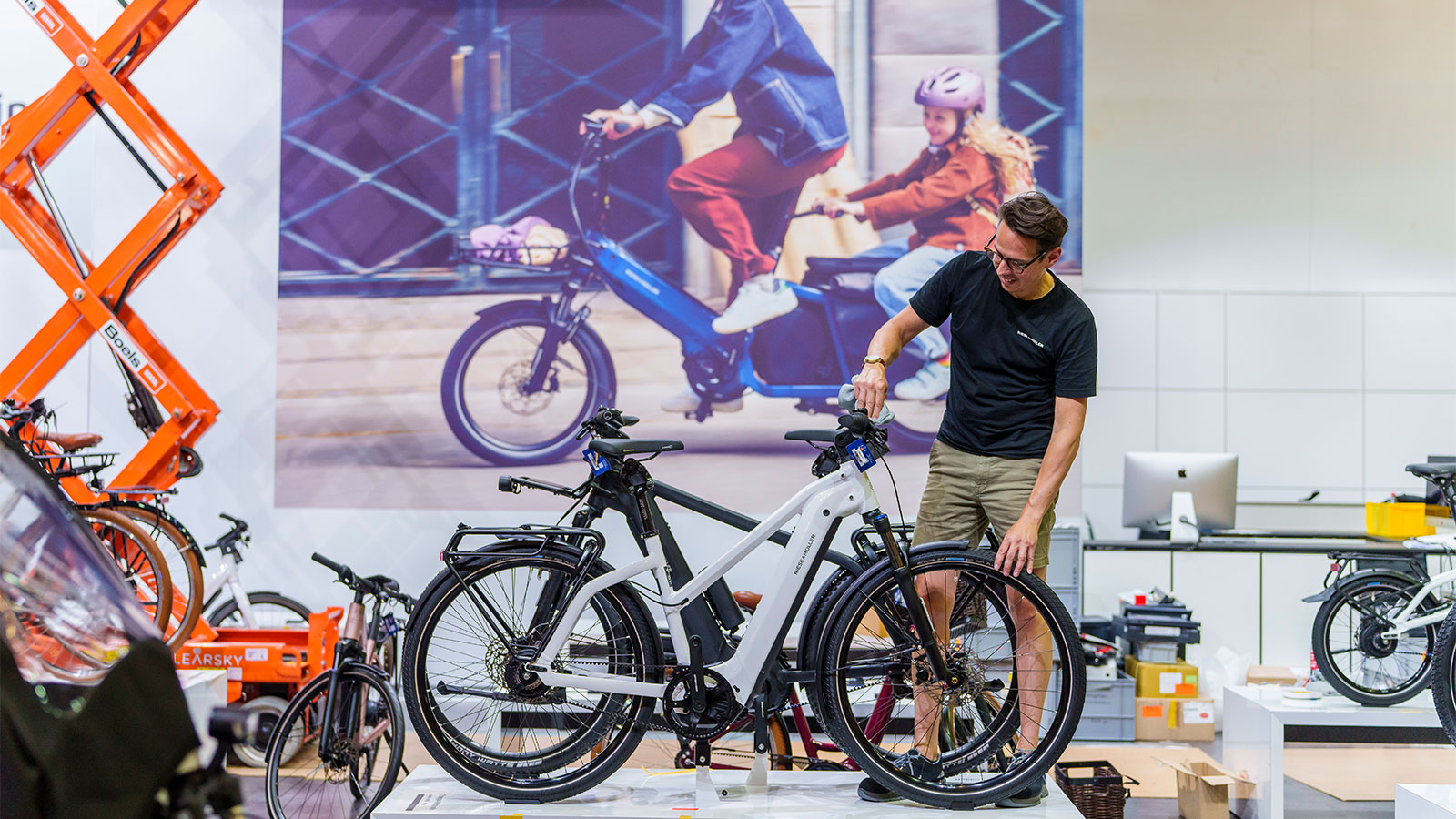 Um homem de camiseta preta fica atrás de uma bicicleta elétrica branca em um pedestal em frente a uma tela que mostra uma bicicleta de carga