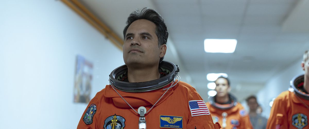 A Million Miles Away'de José M. Hernández rolünde Michael Peña, turuncu bir uzay giysisiyle koridorda yürüyor.