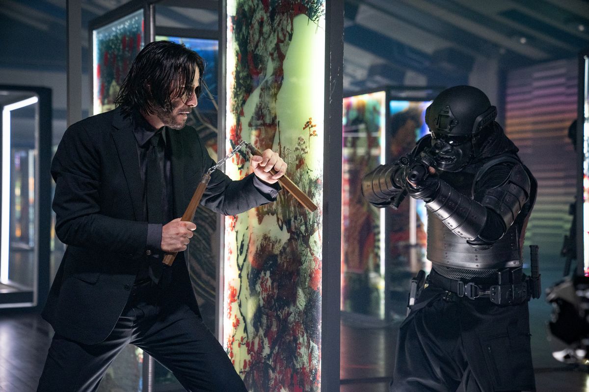 John Wick: Chapter 4'te siyah samuray zırhlı bir askeri pusuya düşürmeye hazırlanan cam bir panelin arkasında num-chuk'ları tutan John Wick rolünde Keanu Reeves.