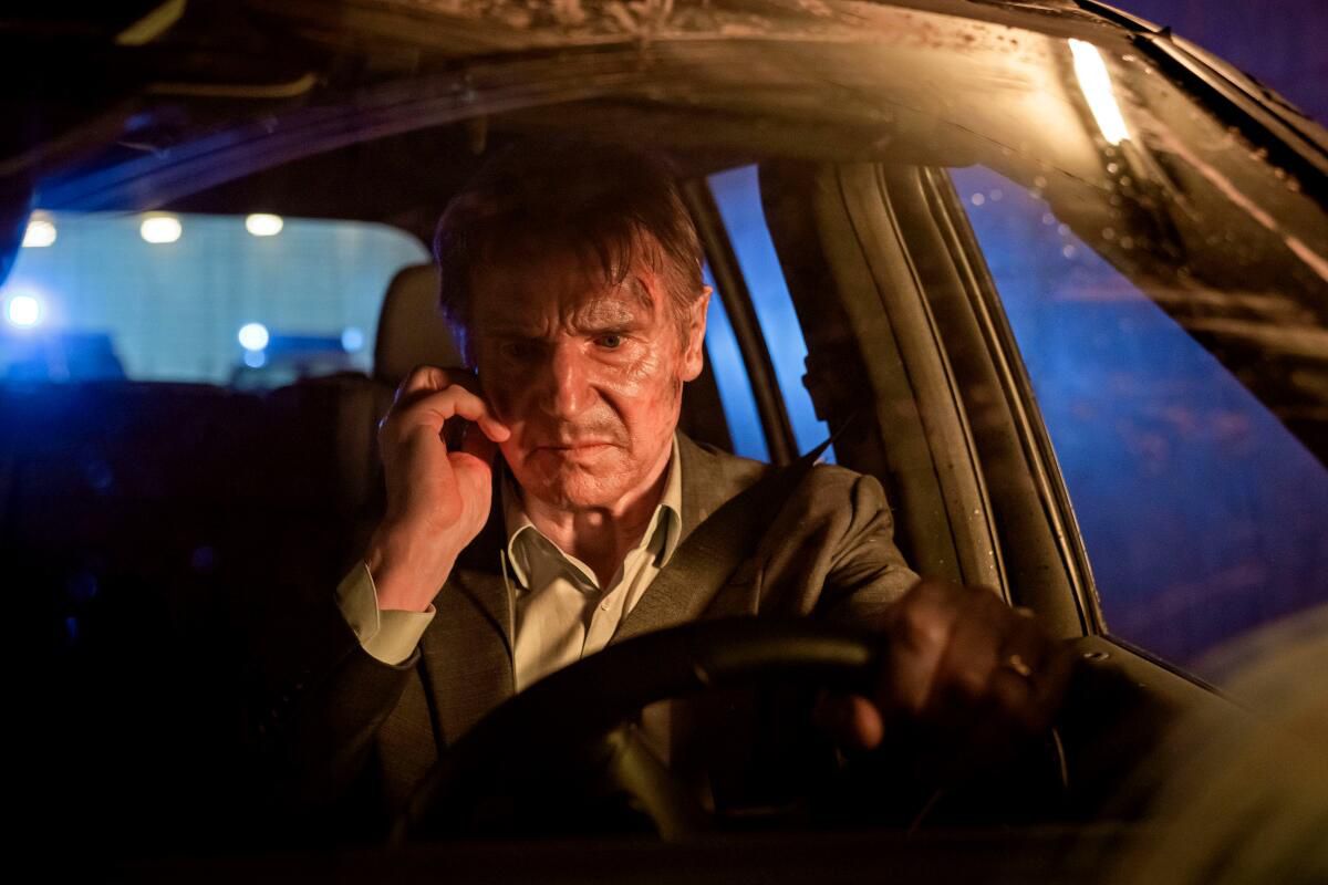 Liam Neeson, zichtbaar slordig, zit achter het stuur van een auto met een mobiele telefoon naast zijn oor in Retribution.