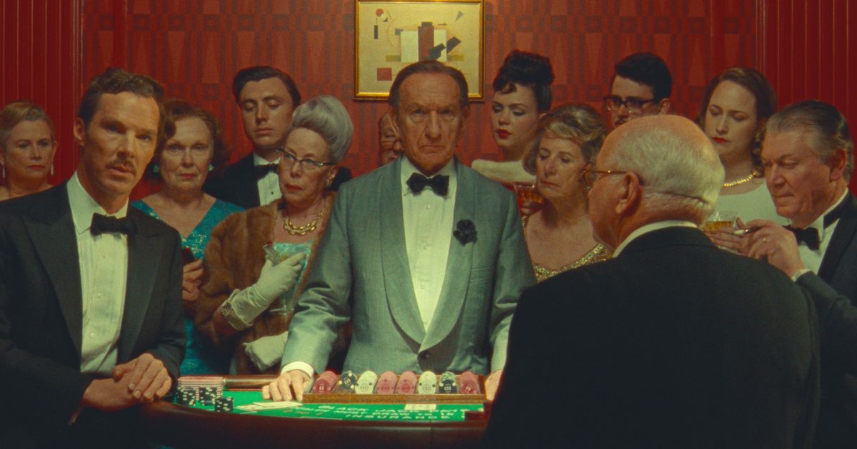 Benedict Cumberbatch (in smoking als Henry Sugar) en Sir Ben Kingsley (als croupier) kijken in de camera terwijl ze aan een tafel in een casino staan, omringd door een nieuwsgierige menigte goedgeklede mensen, in Wes Andersons Netflix-film The Prachtig verhaal van Henry Sugar