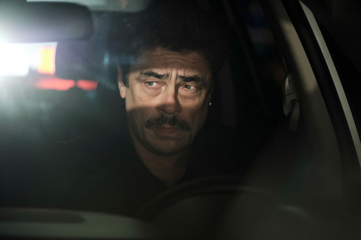 『爬虫類』で夜に車のハンドルの後ろに座り、後部の窓からヘッドライトを照らしているトム・ニコルズを演じるベニチオ・デル・トロ。