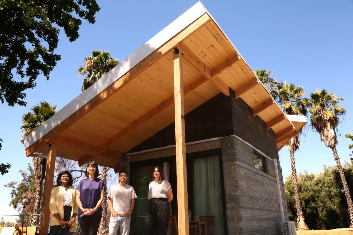 Cuatro personas se encuentran en el porche delantero de una pequeña casa de hormigón con un espectacular techo inclinado de madera.