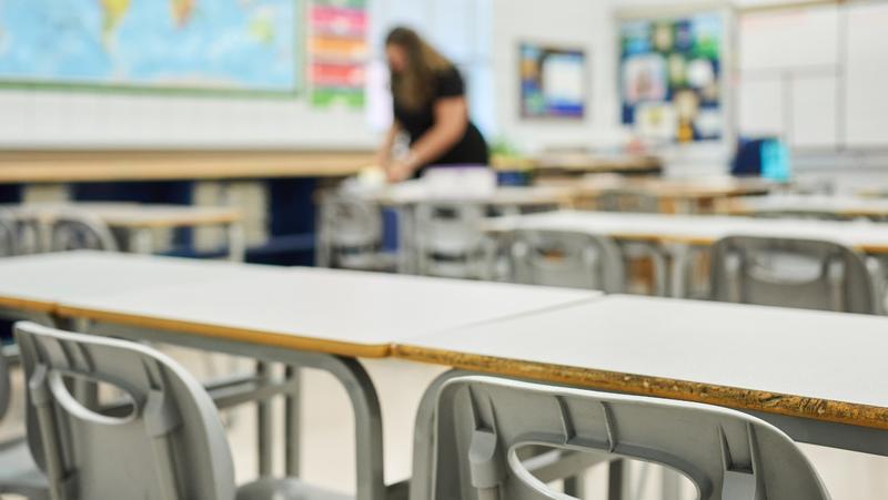 Stoelen en bureaus in een klaslokaal in focus met een leraar op de onscherpe achtergrond, foto door AJ_Watt/Getty Images