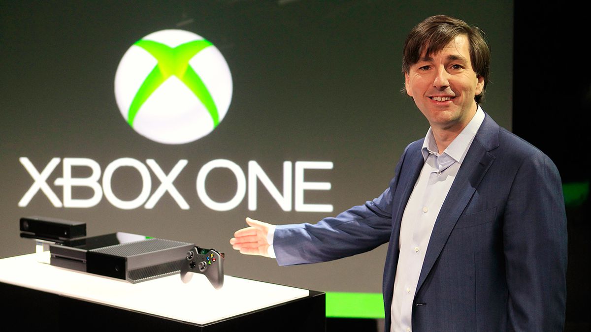 Een directeur met slap haar, Don Mattrick, glimlacht terwijl hij naar een Xbox One-console gebaart voor een groot Xbox One-logo