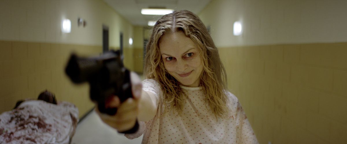 Elizabeth (Heather Graham) peker en pistol inn i kameraet med et forferdelig smil der hun står i en gulnende sykehusgang i en sykehuskåpe i Suitable Flesh
