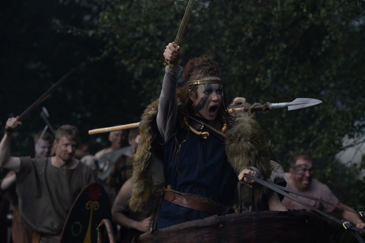 أولغا كوريلينكو، بطلاء وجهها باللون الأزرق، تحمل رمحًا وتصرخ في عربة بينما يقف المحاربون خلفها في بوديكا.