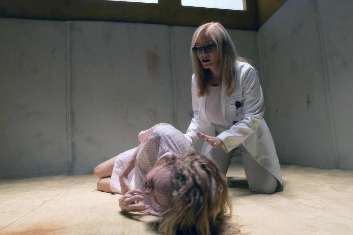 Elizabeth (Heather Graham, con una bata de hospital), se acurruca llorando en el suelo de una celda desnuda de un hospital psiquiátrico mientras una amiga psiquiatra (Barbara Crampton) se arrodilla junto a ella y le tiende una mano solidaria en Adecuado carne