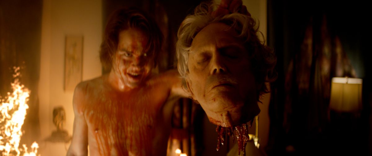 Asa (Judah Lewis), shirtless, drenched in blood, and grinning unnervingly, stands in a house with a fire creeping up the wall behind him and holds up a severed head dripping with gore in Suitable Flesh