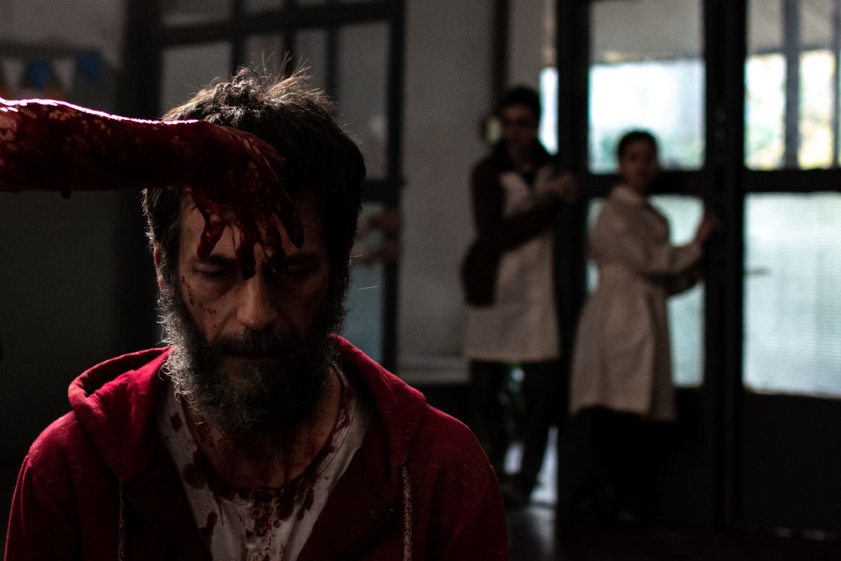 A blood-covered man (Ezequiel Rodríguez) in a red hoodie, head bowed, kneels in the foreground as a mostly offscreen figure places a blood-drenched hand on his forehead. Out-of-focus figures stand behind him, up against a series of glass doors, in When Evil Lurks