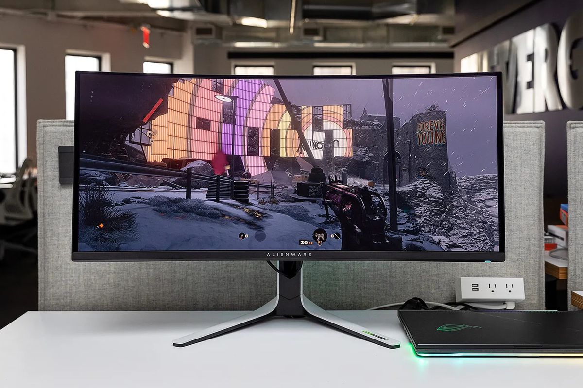 흰색 책상 위에 게임용 노트북에 연결된 Alienware QD-OLED AW3423DW 곡선형 게임 모니터 사진. Deathloop 게임이 화면에 표시되고 있습니다.
