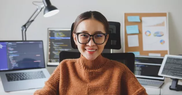 Chân dung lập trình viên trẻ, kỹ sư phần mềm, nhân viên hỗ trợ CNTT, đeo kính nhìn vào camera và mỉm cười thích thú khi làm việc tại nhà.
