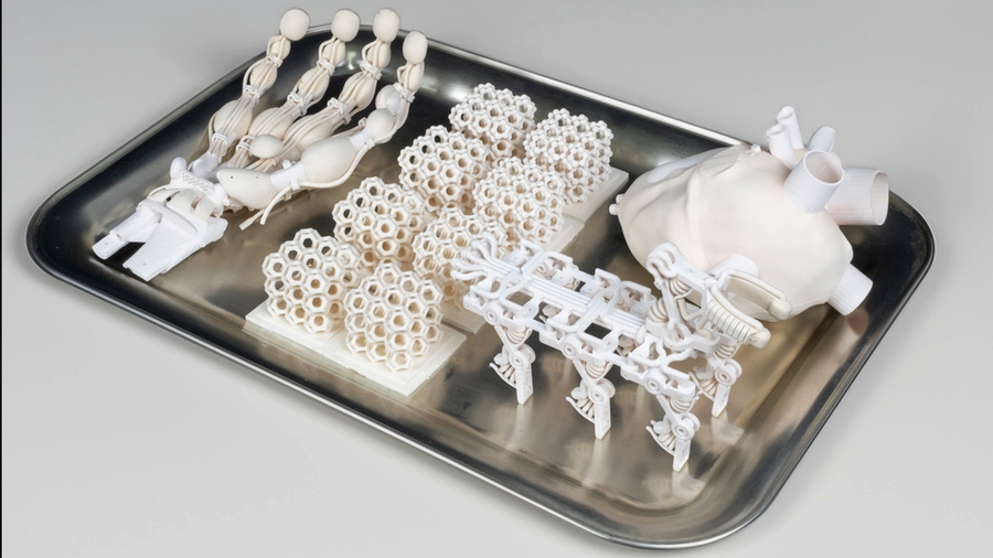 A photograph shows a variety of 3D-printed objects in white, displayed on a tray