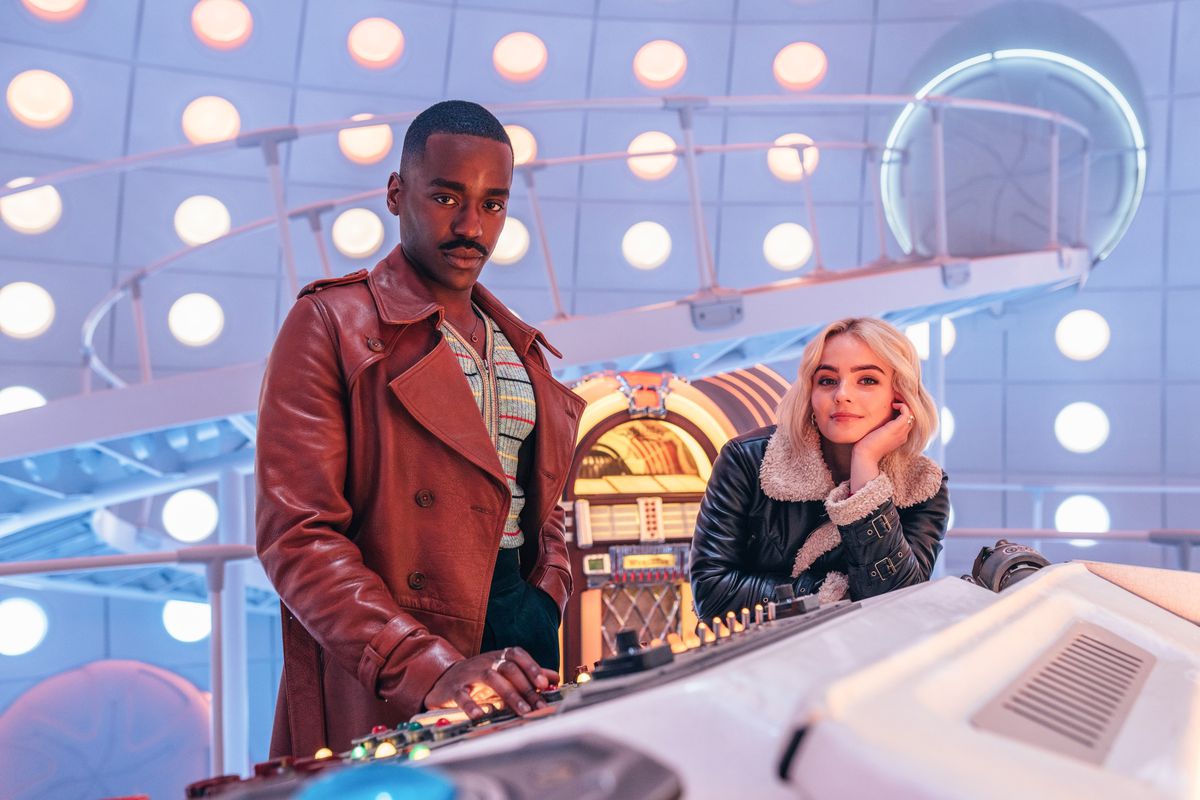 De Dokter poseert vriendelijk achter de knoppen van zijn TARDIS terwijl Ruby Sunday op de console leunt en grijnst in een afbeelding uit de Doctor Who Christmas-special.
