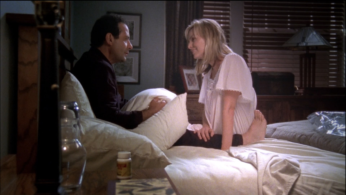 Monk (Tony Shalhoub) sostiene la almohada de Trudy y parece triste mientras su fantasma (Melora Hardin) se sienta frente a él sonriendo.