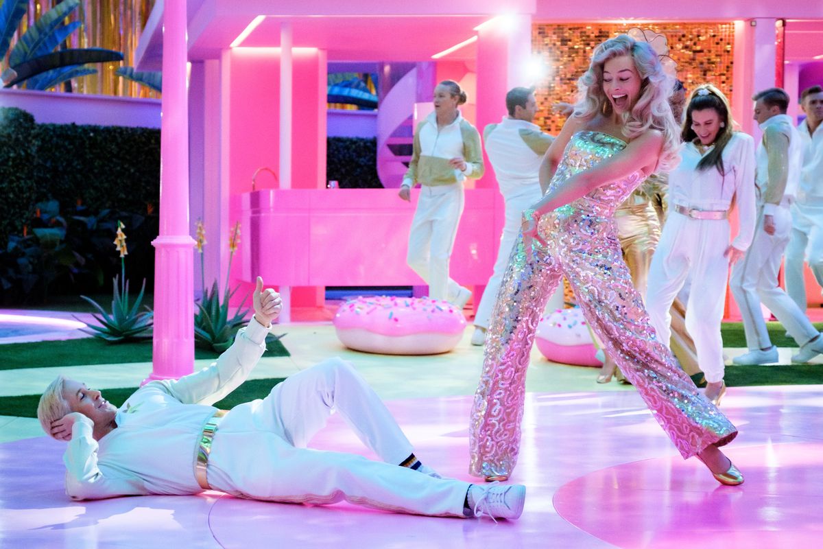 Barbie (Margot Robbie), in een zilverkleurige jumpsuit, grijnst naar Ken (Ryan Gosling), in een geheel witte outfit, liggend op een glanzend roze dansvloer met andere dansende Barbies en Kens op de achtergrond in Greta Gerwig's live-action 2023 film Barbie
