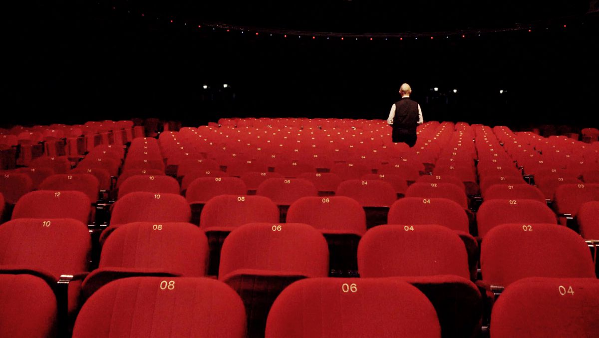 Robert Fripp, der eine Weste trägt, steht mit dem Rücken zur Kamera und ist von leeren roten Zuschauerstühlen umgeben in „Im Hof ​​des purpurnen Königs: King Crimson at 50“.