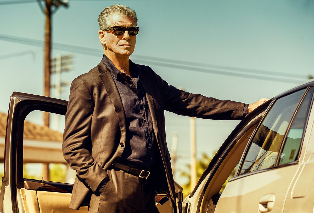 Pierce Brosnan lehnt in Anzug und Sonnenbrille an einem Auto mit offener Tür in „Fast Charlie“.