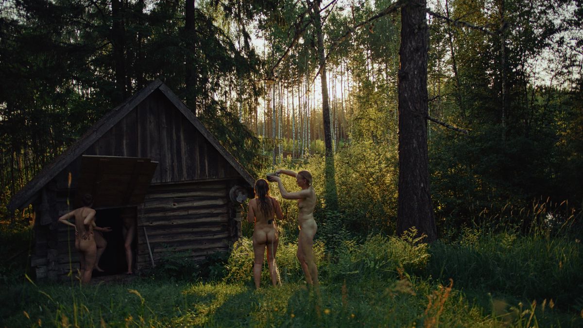 Cuatro mujeres desnudas se encuentran alrededor o dentro de una pequeña sauna hecha de troncos crudos, de pie en un bosque vívidamente verde, en Smoke Sauna Sisterhood.
