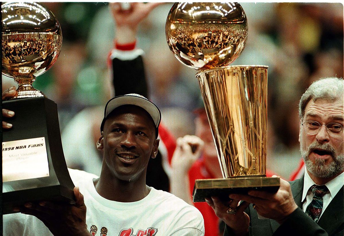 마이클 조던은 MVP 결승전 트로피를 들고 있고, 필 잭슨은 1998년 NBA 챔피언십 트로피를 들고 있습니다.