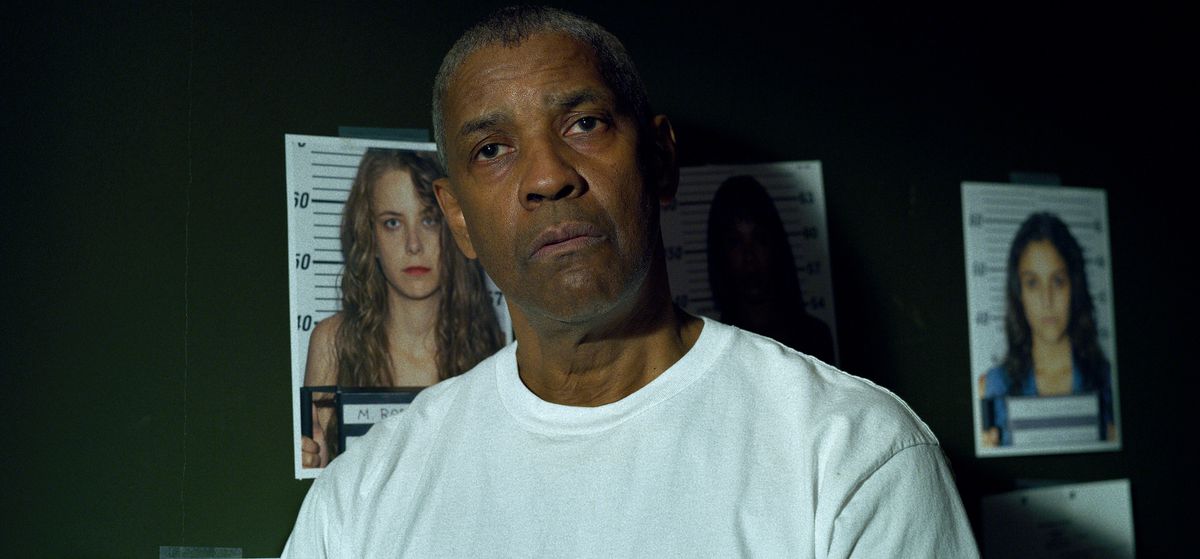 Denzel Washington은 흰색 티셔츠를 입고 The Little Things에서 살해된 여성의 사진이 있는 녹색 벽 앞에 서 있습니다.