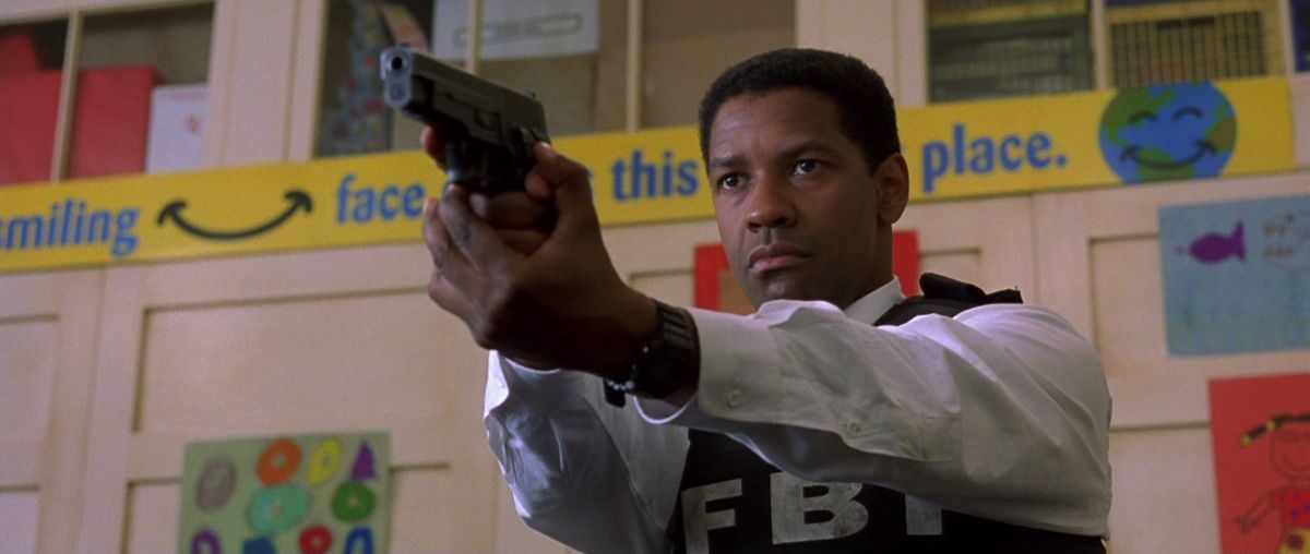 Denzel Washington usa un chaleco antibalas que dice "FBI" y apunta con un arma mientras está parado en lo que parece el gimnasio de una escuela en The Siege.