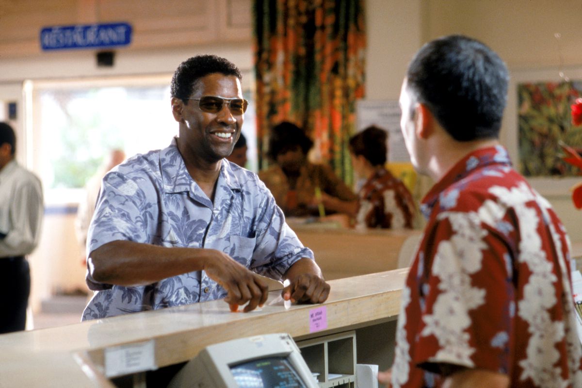 دينزل واشنطن مبتسمًا، يرتدي قميص هاواي، ويسجل وصوله إلى فندق في منطقة Out of Time.