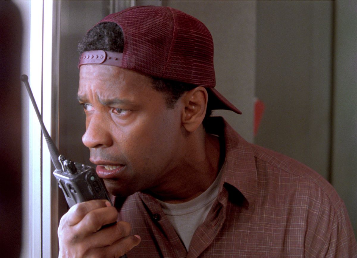 Denzel Washington, wearing a backwards baseball hat and a collared shirt, talks on a walkie-talkie while looking out of a window in John Q.