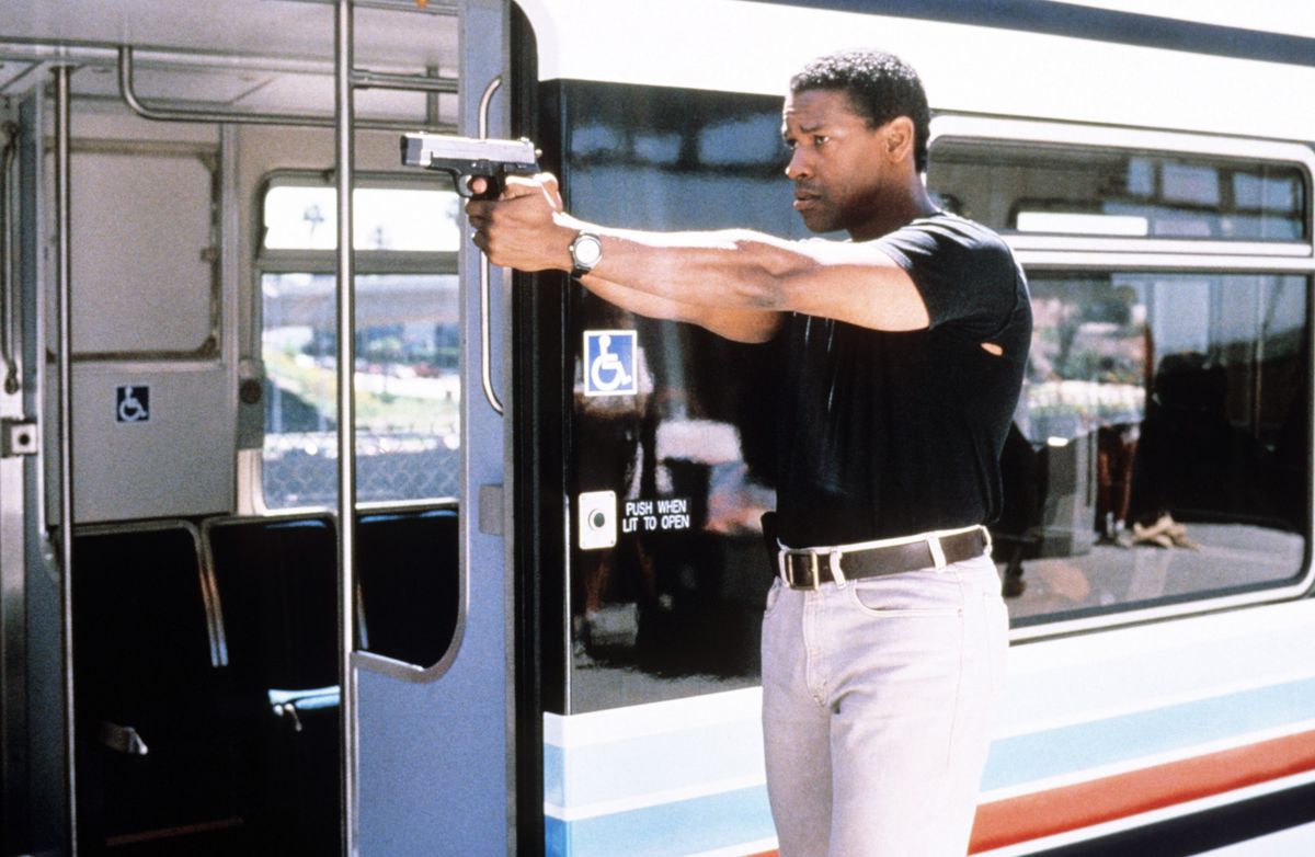 Denzel Washington, gekleed in een strak zwart t-shirt, houdt een pistool vast naast een treinwagon in Virtuosity.