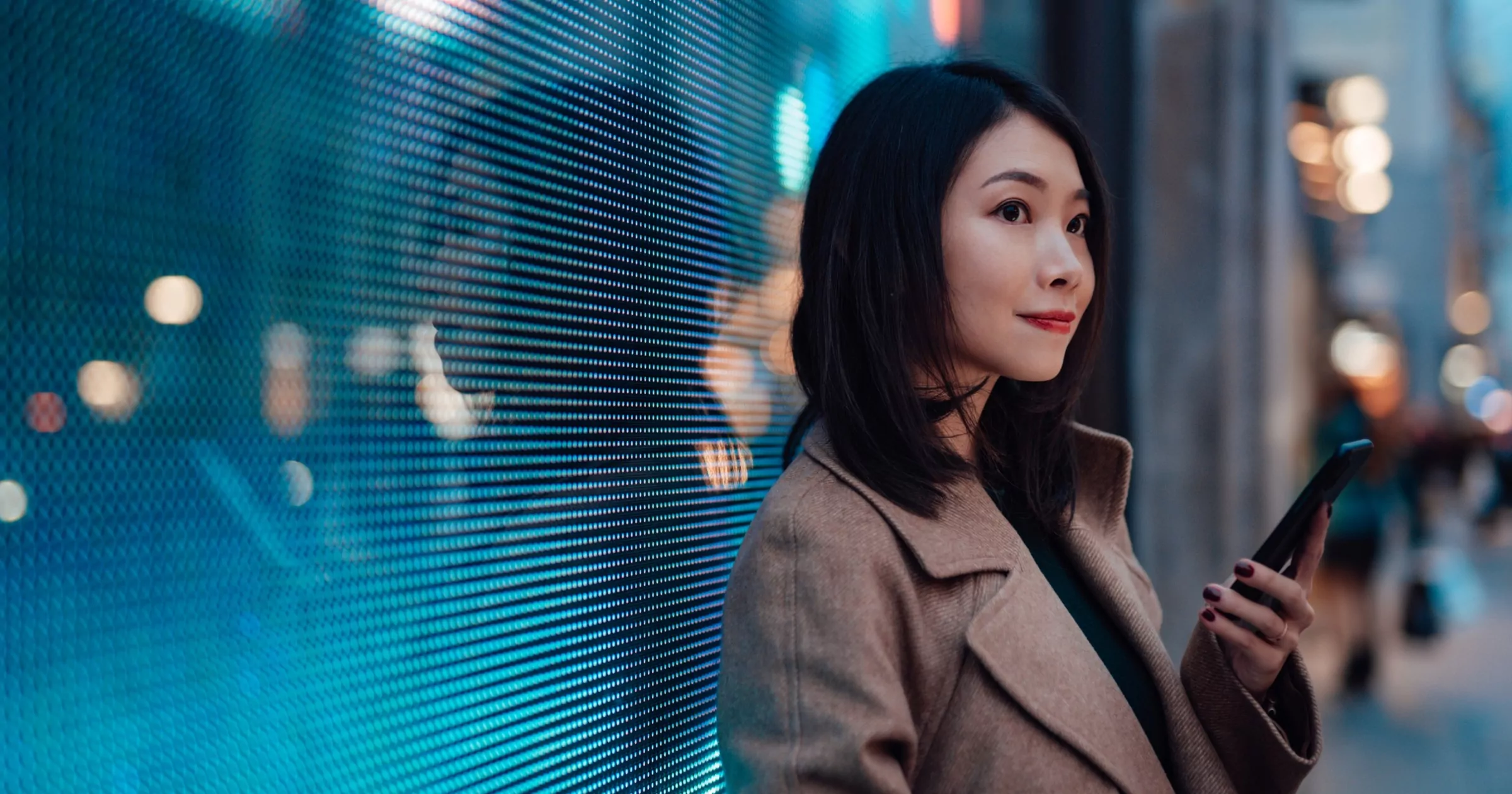 Geceleri şehir caddesinde beklerken cep telefonu kullanan genç Asyalı iş kadını. Arka planda şehir ışıklarının aydınlatıldığı devasa bir dijital ekranın önünde. Geleceği bağlayın.