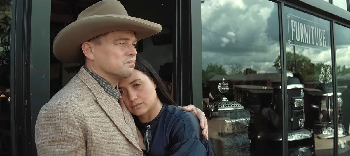 Ernest (Leonardo DiCaprio, ruskea puku ja cowboy-hattu) lohduttaa vaimoaan Molliea (Lily Gladstone), kun tämä nojautuu häntä vasten huonekaluliikkeen ikkunan edessä Killers of the Flower Moonissa.