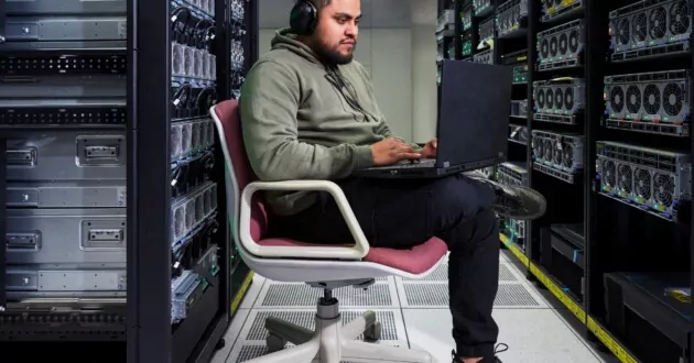 Person sitzt auf einem Stuhl in einem Datenspeicher, trägt Kopfhörer und arbeitet am Laptop